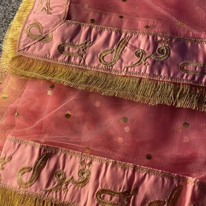 May include: A pink sheer fabric with gold embroidered detailing and fringe. The fabric has a dotted pattern and a satin border with ornate gold embroidery. The edges are trimmed with gold fringe.