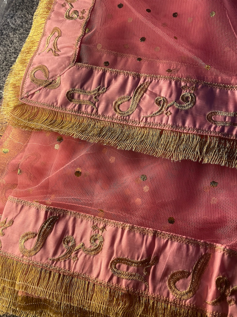 May include: A pink sheer fabric with gold embroidered detailing and fringe. The fabric has a dotted pattern and a satin border with ornate gold embroidery. The edges are trimmed with gold fringe.
