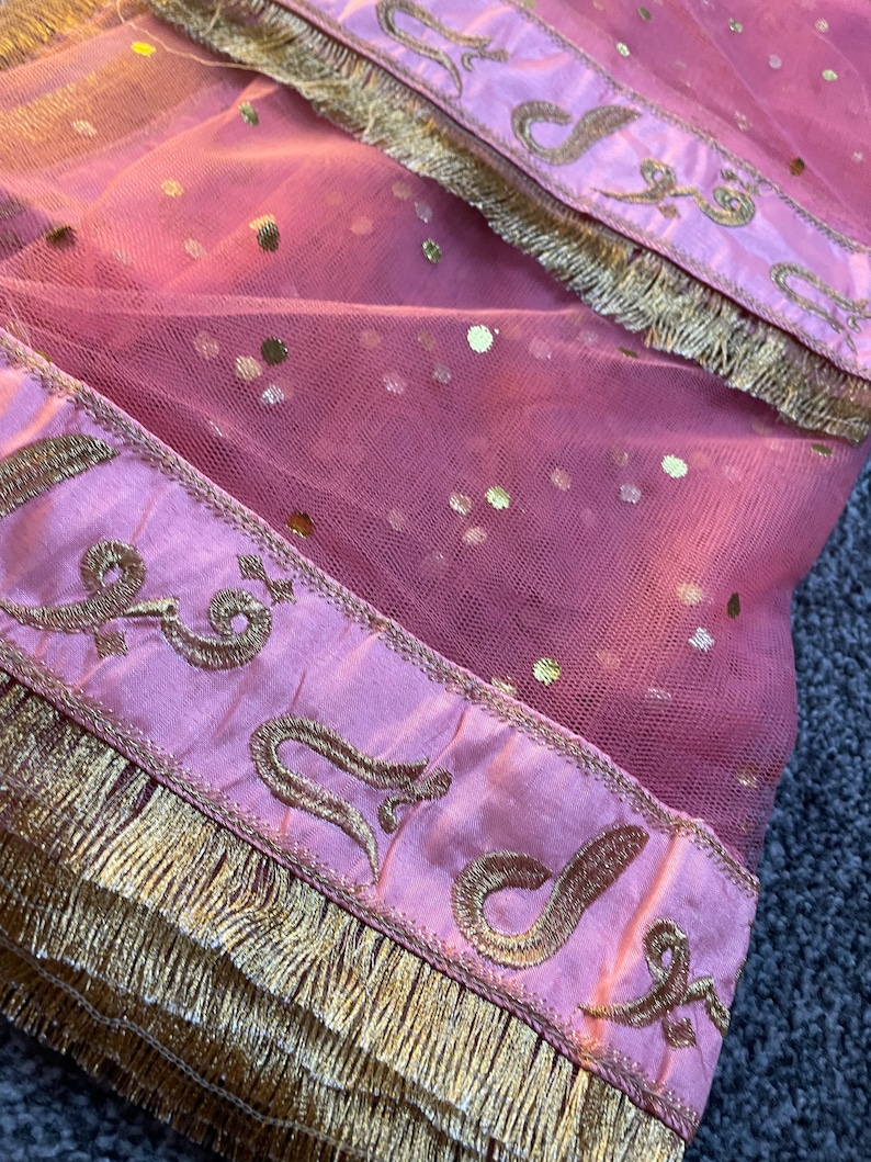 May include: A pink net fabric with gold sequins and decorative borders. The borders are a lighter pink with gold embroidery and a gold fringe. The fabric is folded, showing the texture and details of the design.