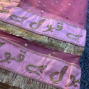 May include: Close-up of a pink sheer fabric with gold embellishments. The fabric has gold dots and is trimmed with a pink border featuring gold embroidery and a gold fringe. The embroidery includes Arabic script.