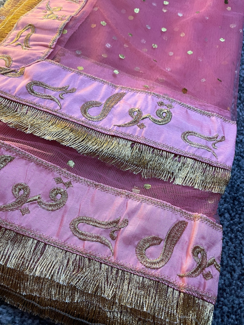 May include: Close-up of a pink sheer fabric with gold embellishments. The fabric has gold dots and is trimmed with a pink border featuring gold embroidery and a gold fringe. The embroidery includes Arabic script.