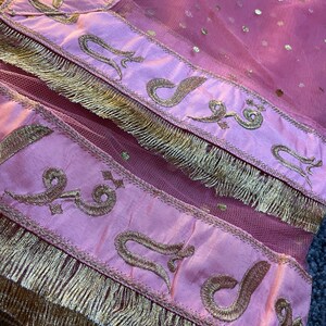 May include: Close-up of a pink textile with gold embroidery and fringe. The fabric has a sheer, dotted overlay. Gold embroidered script is featured on a pink satin border. The bottom edge is trimmed with gold fringe.
