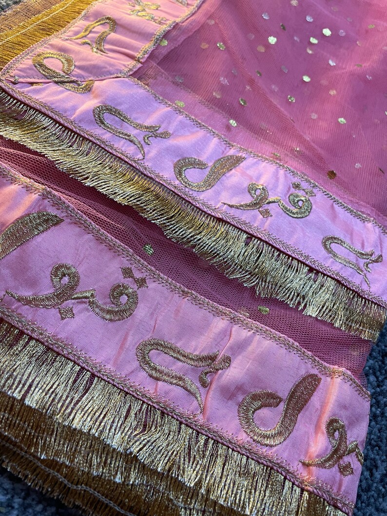 May include: Close-up of a pink textile with gold embroidery and fringe. The fabric has a sheer, dotted overlay. Gold embroidered script is featured on a pink satin border. The bottom edge is trimmed with gold fringe.