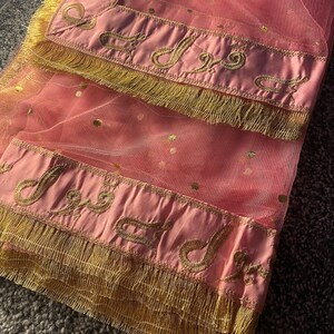 May include: A pink net scarf with gold polka dots and a satin border. The border is embroidered with gold script and trimmed with gold fringe. The scarf is folded, showing the details of the design.