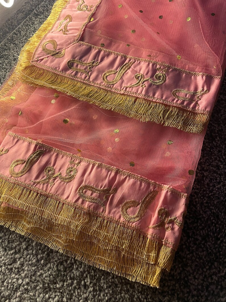 May include: A pink net scarf with gold polka dots and a satin border. The border is embroidered with gold script and trimmed with gold fringe. The scarf is folded, showing the details of the design.