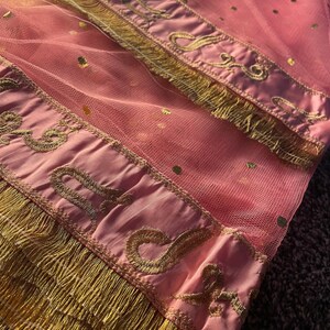 May include: Pink tulle fabric with gold polka dots and gold embroidered trim. The trim features an ornate design and is finished with a gold fringe. The fabric is folded over, showing the details of the design.