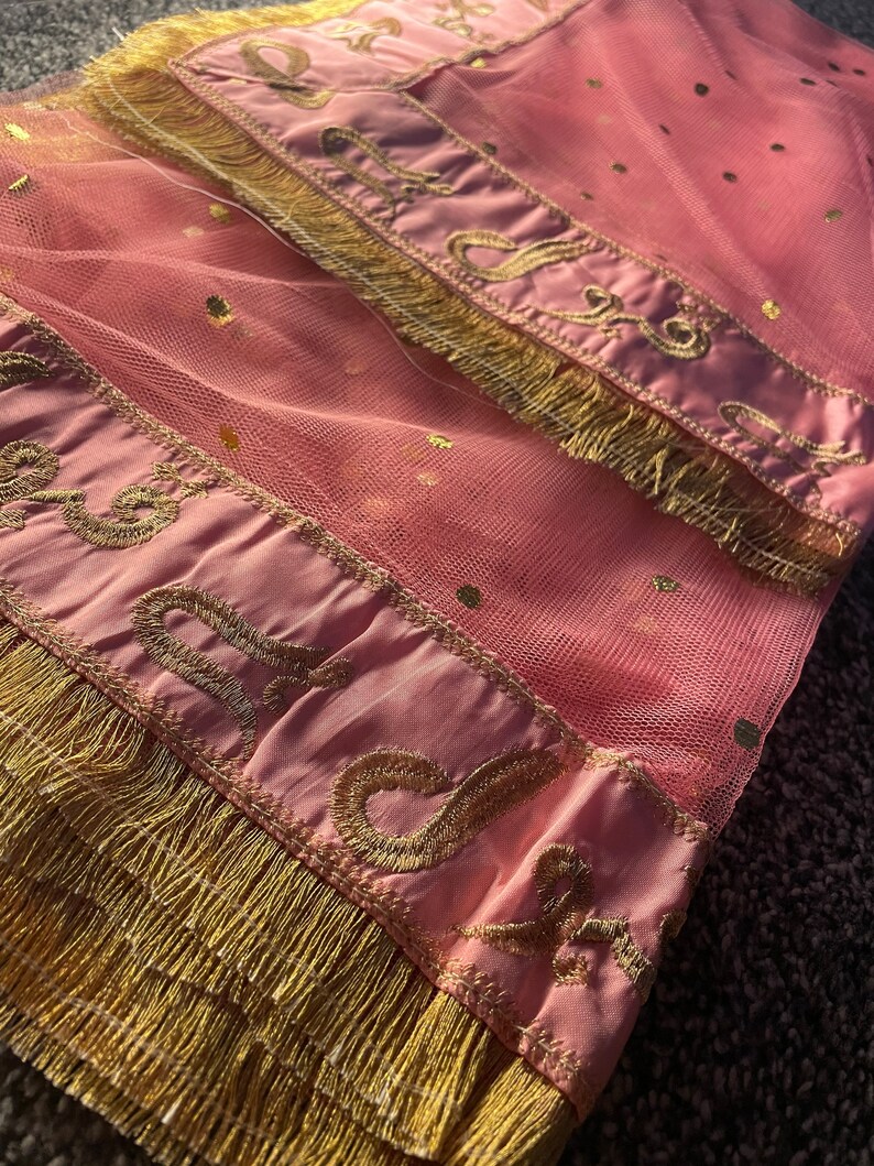 May include: Pink tulle fabric with gold polka dots and gold embroidered trim. The trim features an ornate design and is finished with a gold fringe. The fabric is folded over, showing the details of the design.