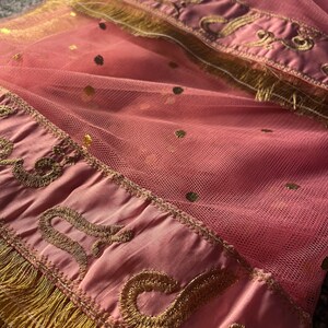 May include: Pink tulle fabric with gold polka dots and a decorative border. The border features gold embroidery and fringe. The fabric is folded, creating a layered effect.