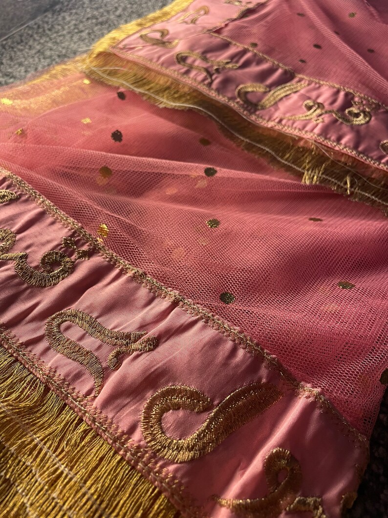 May include: Pink tulle fabric with gold polka dots and a decorative border. The border features gold embroidery and fringe. The fabric is folded, creating a layered effect.