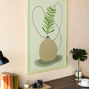 May include: A framed print of a stylized green plant in a brown vase with a red line abstract design on a light green background. The print is hanging on a light beige wall above a wooden desk with a camera, books, a small potted plant, a laptop, a glass of water, and a white coffee cup.