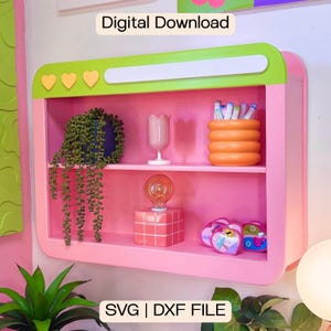Puede incluir: Una estantería de pared rosa con un borde superior verde lima que presenta tres iconos de corazón amarillos y una barra rectangular blanca. El texto Digital Download y SVG | DXF FILE es visible. La estantería contiene una planta colgante, una copa rosa, un recipiente naranja acanalado con marcadores, una bombilla sobre una base de rejilla rosa y pequeños objetos en forma de corazón.