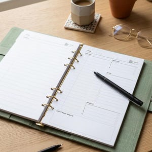 Peut inclure: Un agenda ouvert de couleur vert sauge avec des anneaux métalliques dorés, présentant des pages blanches avec des sections imprimées pour l'agenda, les priorités, les listes de tâches et les routines. Un stylo noir repose sur la page.