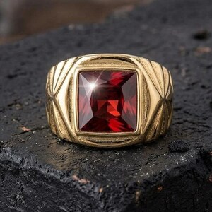Puede incluir: Un anillo de tono dorado con una gran piedra preciosa de color rojo intenso y corte cuadrado en el centro. La banda presenta un diseño ancho y texturizado con patrones geométricos angulares que irradian desde el engaste central, creando un aspecto audaz.