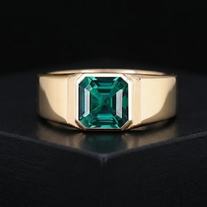 Könnte beinhalten: Ein goldener Ring mit einem rechteckigen, smaragdgeschliffenen grünen Edelstein. Der Ring hat ein dickes Band und ein schlichtes, modernes Design. Der Edelstein ist in einer quadratischen Fassung gefasst.