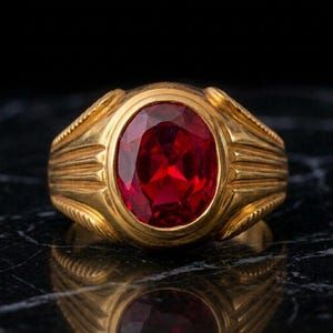 Puede incluir: Un anillo de oro con una gran piedra preciosa de color rojo intenso y corte ovalado en el centro. La banda del anillo presenta un diseño texturizado y acanalado que se extiende desde el engaste central, creando una apariencia estructurada.