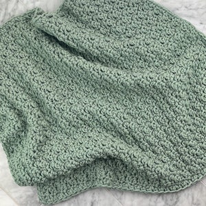 May include: A soft, sage green crocheted blanket with a textured stitch pattern. The blanket is folded and draped over a marble surface.
