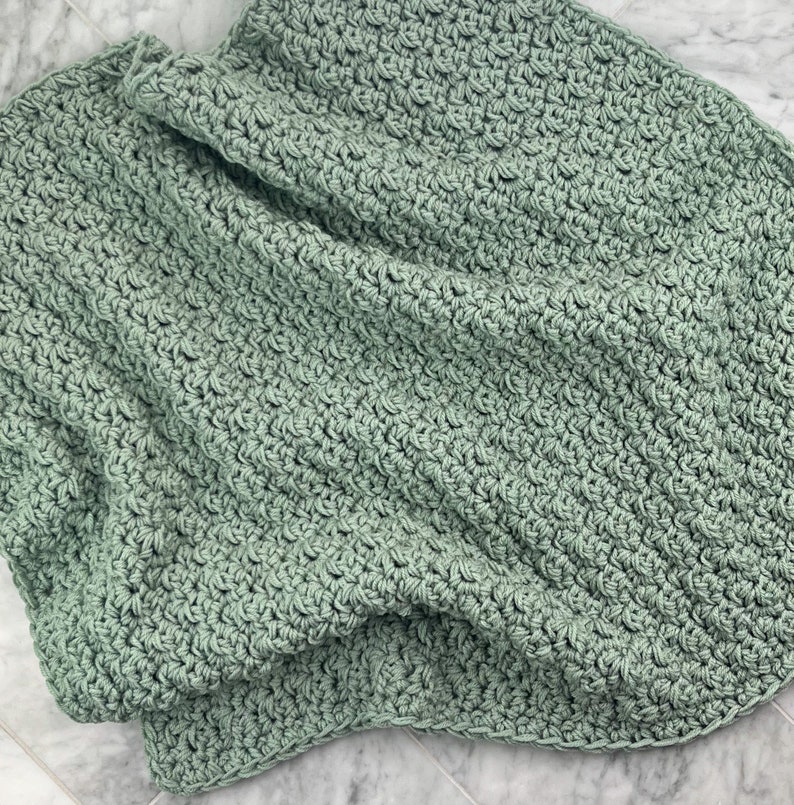 May include: A soft, sage green crocheted blanket with a textured stitch pattern. The blanket is folded and draped over a marble surface.