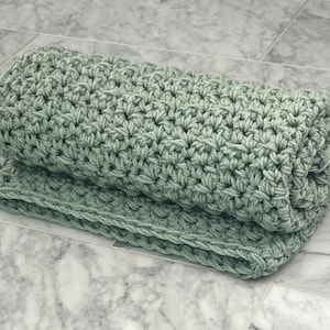 May include: A sage green crocheted blanket rolled up on a marble surface.