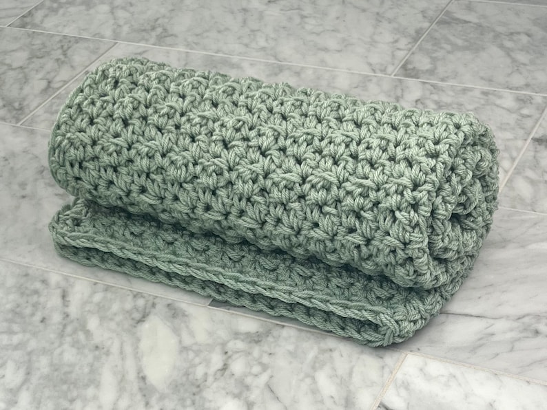 May include: A sage green crocheted blanket rolled up on a marble surface.