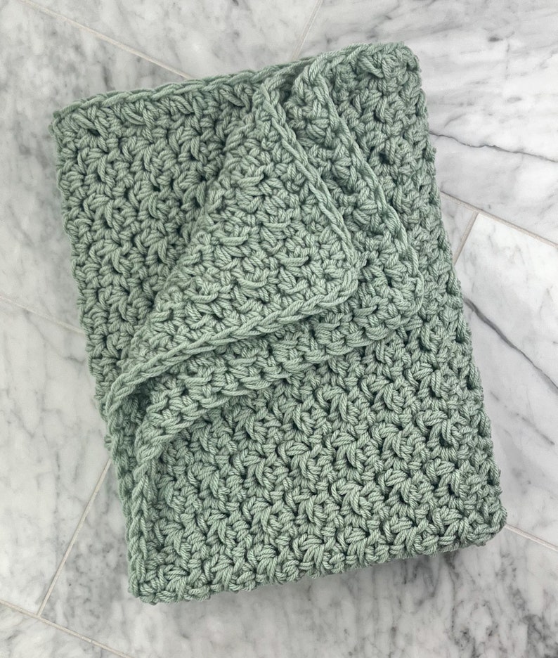 May include: A soft, sage green crocheted blanket with a textured stitch pattern. The blanket is folded in half and the edges are visible.