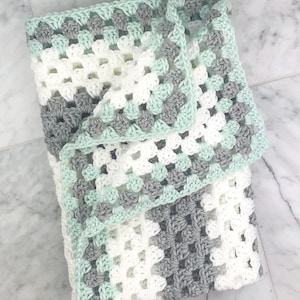 May include: A crocheted baby blanket in white, gray, and light green. The blanket is made with a granny square pattern.