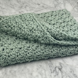 May include: A soft, sage green crocheted blanket folded on a marble surface.