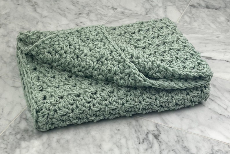May include: A soft, sage green crocheted blanket folded on a marble surface.