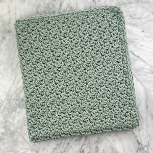 May include: A soft, sage green crocheted blanket with a simple stitch pattern. The blanket is folded and lying on a white marble surface.