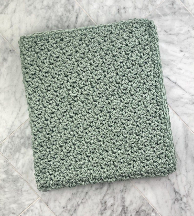 May include: A soft, sage green crocheted blanket with a simple stitch pattern. The blanket is folded and lying on a white marble surface.