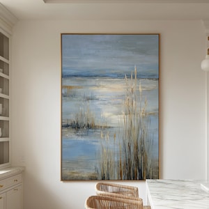 May include: An abstract landscape painting with a gold frame. The artwork features a serene scene with blue, beige, and gray tones, depicting water and tall grasses. Wicker bar stools with black legs are in the foreground.
