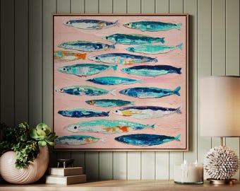 Op de afbeelding: Een olieverfschilderij op canvas toont een school vissen in tinten blauw, turquoise en oranje tegen een roze achtergrond. Het kunstwerk is ingelijst in een lichtbruine houten lijst en wordt tentoongesteld op een houten kast.