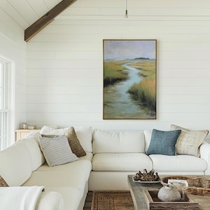 May include: A living room with a white sectional sofa, throw pillows, and a wooden coffee table. A framed landscape painting hangs on the white shiplap wall. The painting depicts a marsh scene with a winding waterway and grassy banks.