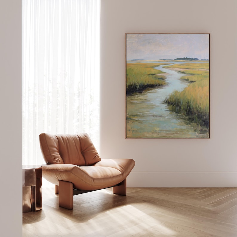 May include: A modern living room featuring a large landscape painting with a gold frame, a brown leather armchair, and a small side table. The painting depicts a marsh scene with a winding river and tall grasses. The room has hardwood floors and a sheer white curtain.
