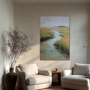 May include: An oil painting of a marsh scene with a winding waterway, framed in a light wood. The artwork features a palette of greens, blues, and yellows, with a muted sky. The painting hangs above two cream-colored armchairs.