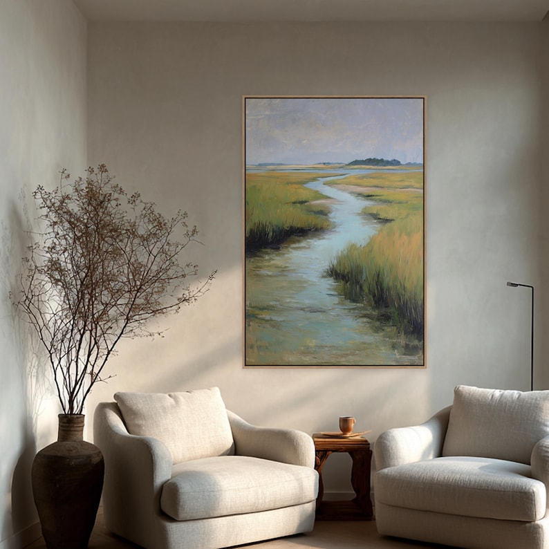 May include: An oil painting of a marsh scene with a winding waterway, framed in a light wood. The artwork features a palette of greens, blues, and yellows, with a muted sky. The painting hangs above two cream-colored armchairs.