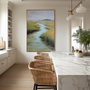 May include: A kitchen scene featuring a large landscape painting with a river winding through marshy grasses. Three woven rattan bar stools with black metal legs are positioned in front of a marble countertop. A white cabinet and a brass light fixture are also visible.