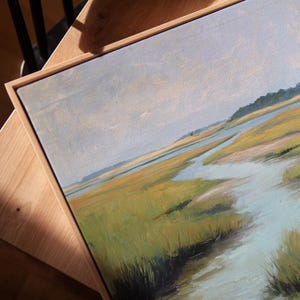 May include: An oil painting of a marsh landscape with a light blue sky. The artwork features a palette of greens, yellows, and blues, with a wooden frame. The painting depicts a serene scene of water and grassy areas.
