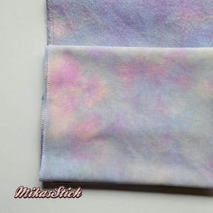 Könnte beinhalten: Zwei Stoffstücke mit einem Batik-Muster in Rosa-, Lila- und Blautönen. Der Stoff ist in der Mitte gefaltet und die Kanten sind vernäht.