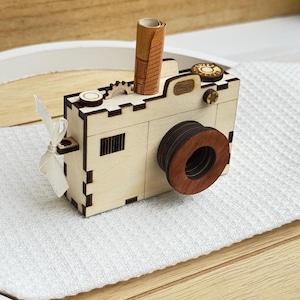 Könnte beinhalten: Ein helles Holzkameramodell mit einem dunkelbraunen, kreisförmigen Objektiv, einem kleinen zahnradförmigen Detail auf der Oberseite und einem aufgerollten Stück Papier, das in den oberen Schlitz gesteckt ist. Ein weißes Stoffband ist an der Seite befestigt.