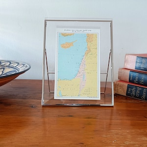 Puede incluir: Un mapa enmarcado de Israel, exhibido en un soporte de acrílico transparente. El mapa presenta un diseño colorido con tonos azules, amarillos y verdes, sobre una superficie de madera. También se ven un cuenco decorativo y libros apilados.