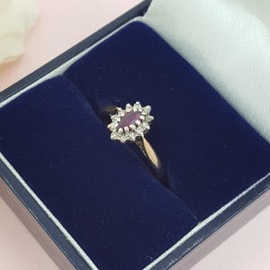 May include: A gold ring featuring a central oval ruby, encircled by smaller, clear gemstones. The ring is presented in a dark blue velvet-lined jewellery box with a white satin interior.