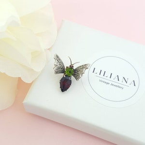 May include: A silver butterfly brooch with a red gemstone and green accents. The brooch is on a white box with the text "Liliana Vintage Jewellery".