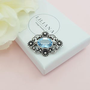 May include: A silver-toned brooch with a large, oval, light blue gemstone at its center. The brooch is surrounded by a decorative floral design. The brooch is on a white box with the text "LILIANA Vintage Jewellery".