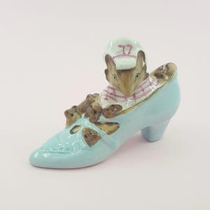 May include: A blue ceramic shoe figurine with a mouse wearing a pink and white striped scarf and a white hat with the number 77 on it. The mouse is sitting in the shoe with other smaller mice figurines.