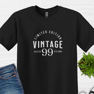 Peut inclure: T-shirt noir avec un texte blanc qui dit "Édition limitée Vintage Âgé de 99 ans". Le t-shirt est présenté sur une surface en bois blanc avec un jean bleu, des lunettes de soleil et une paire de baskets blanches.
