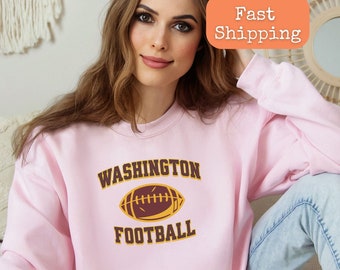 Op de afbeelding: Lichtroze sweatshirt met de tekst "WASHINGTON FOOTBALL" en een bruine en gele voetbalafbeelding.