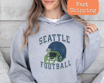 Könnte beinhalten: Ein grauer Kapuzenpullover mit dem Text "SEATTLE FOOTBALL" und einer Grafik eines Vintage-Footballhelms.