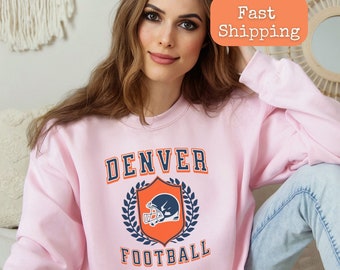 May include: Light pink sweatshirt with a navy blue and orange graphic of a football helmet with the text "DENVER FOOTBALL" in a vintage collegiate style.