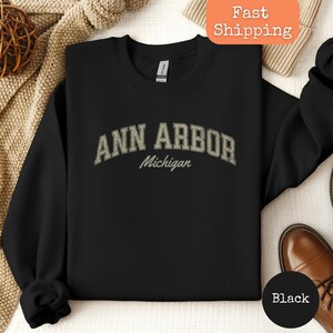 Könnte beinhalten: Schwarzes Sweatshirt mit Rundhalsausschnitt und dem Text "ANN ARBOR Michigan" in einem Vintage-Schriftstil. Das Sweatshirt besteht aus einem weichen Material. Ein klassischer Stil, ein tolles Geschenk für alle, die Ann Arbor, Michigan lieben.