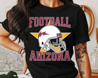 Op de afbeelding: Zwart T-shirt met een witte voetbalhelm en een gele ster. De tekst "FOOTBALL ARIZONA" is in maroon op het shirt gedrukt.